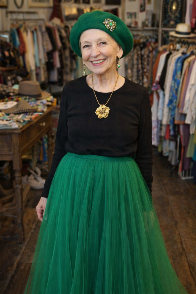 A petite 75 year old woman wearing a green beret, no hair showing. The beret has a broach pinned to it. Dangling earrings, a gold neclace with a flower pendant, a black long sleeve tee shirt, and a long flowing green tulle skirt. (AI produced.) New Orleans