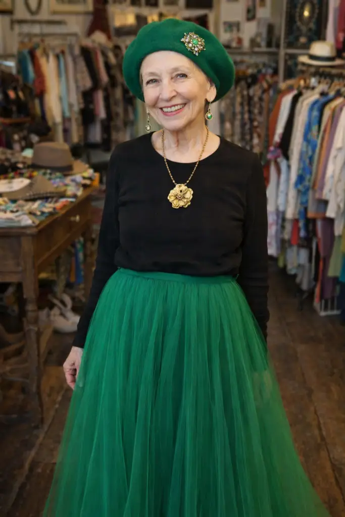 A petite 75 year old woman wearing a green beret, no hair showing. The beret has a broach pinned to it. Dangling earrings, a gold neclace with a flower pendant, a black long sleeve tee shirt, and a long flowing green tulle skirt. (AI produced.) New Orleans