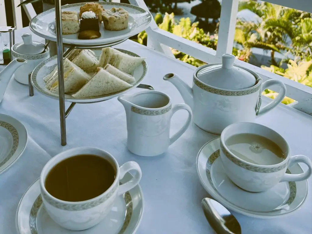 Two cups, a white teapot, and a tiered tray of small sandwiches and pastries on a white tablecloth beside a porch railing.