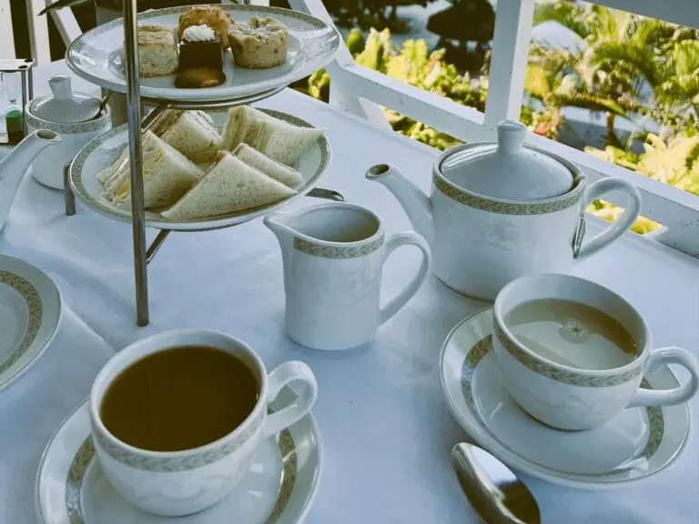 Two cups, a white teapot, and a tiered tray of small sandwiches and pastries on a white tablecloth beside a porch railing.